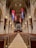 A grand, expansive church interior features rows of wooden pews flanking a long central aisle leading towards the altar. Tall, arched ceilings are adorned with hanging banners in blue, red, and gold hues. Large, ornate chandeliers illuminate the space with a warm glow, while intricate stone architecture displays elegant columns and details.