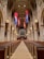 A grand, expansive church interior features rows of wooden pews flanking a long central aisle leading towards the altar. Tall, arched ceilings are adorned with hanging banners in blue, red, and gold hues. Large, ornate chandeliers illuminate the space with a warm glow, while intricate stone architecture displays elegant columns and details.