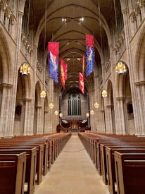 A grand, expansive church interior features rows of wooden pews flanking a long central aisle leading towards the altar. Tall, arched ceilings are adorned with hanging banners in blue, red, and gold hues. Large, ornate chandeliers illuminate the space with a warm glow, while intricate stone architecture displays elegant columns and details.
