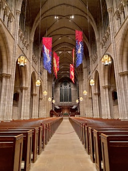 A grand, expansive church interior features rows of wooden pews flanking a long central aisle leading towards the altar. Tall, arched ceilings are adorned with hanging banners in blue, red, and gold hues. Large, ornate chandeliers illuminate the space with a warm glow, while intricate stone architecture displays elegant columns and details.
