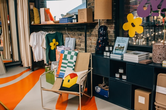 A cozy corner of a modern store featuring a variety of items and decor. There is a chair with a pillow adorned with a colorful house design next to a stack of textiles. A small shelf displays books, candles, and decorative items against a brick wall. Clothing items, including a shirt with a floral logo, hang from a rack. Bright, abstract patterns are painted on the floor, and strings of lights can be seen outside the window, adding to the ambiance.