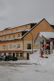 A snow-covered street with a multi-story building featuring beige walls and a dark roof. In front of the building, there is a parked black car labeled 'Carabinieri'. Italian flags are displayed on a nearby railing. The building has multiple windows and signs hanging above its entrance.