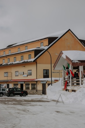 A snow-covered street with a multi-story building featuring beige walls and a dark roof. In front of the building, there is a parked black car labeled 'Carabinieri'. Italian flags are displayed on a nearby railing. The building has multiple windows and signs hanging above its entrance.