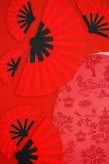 A collection of vibrant red and black patterned fans is arranged against a red background. Each fan is decorated with a black radial pattern emanating from a central point. Beside the fans, there is a red panel featuring intricate designs of pagodas, trees, and birds.