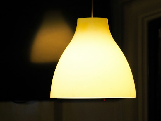 A large, softly glowing pendant lamp with a smooth, matte finish hangs against a dark background. The light emanates a warm, yellowish hue, creating a cozy and inviting ambiance. Shadows and a soft glow of light are visible on the surrounding walls.