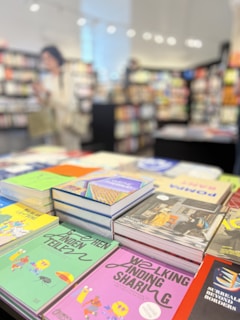 A stack of books on a display table in a bookstore, with various colorful book covers visible. The background is blurred, showing shelves filled with books and a person browsing.