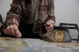 A person dressed in a brown plaid shirt seems to be engaged in a board game. Their hand is reaching towards the game pieces on the table. The game board is partially visible, featuring intricate designs and a noticeable circular emblem with artistic details. Several chairs are seen in the background.