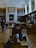The scene is set in a large, ornate library or museum room with high ceilings and shelves filled with books. People are sitting and walking around, engaged with their phones or interacting with the exhibits. The room is illuminated by natural light streaming in through large windows.