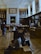 The scene is set in a large, ornate library or museum room with high ceilings and shelves filled with books. People are sitting and walking around, engaged with their phones or interacting with the exhibits. The room is illuminated by natural light streaming in through large windows.