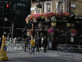 A bustling urban scene featuring a traditional pub adorned with vibrant hanging flowers. People are walking on the sidewalk near the entrance. A red traffic light is visible, along with street signs and metal barricades. The atmosphere suggests a busy city area with a mix of modern and historical elements.