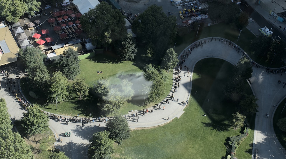 Aerial view of a park area with a large crowd of people forming a long, curved line on a pathway. The park is surrounded by trees and some buildings with red rooftops. The weather appears sunny, casting shadows from the trees and structures onto the greenery and pathway.