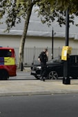 A person in dark clothing, possibly a security or law enforcement officer, is standing next to a black car parked by the roadside. A bright red and yellow vehicle, possibly an emergency service vehicle, is visible nearby. Trees and a metal fence are in the background.