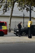 A person in dark clothing, possibly a security or law enforcement officer, is standing next to a black car parked by the roadside. A bright red and yellow vehicle, possibly an emergency service vehicle, is visible nearby. Trees and a metal fence are in the background.