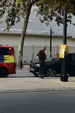 A person in dark clothing, possibly a security or law enforcement officer, is standing next to a black car parked by the roadside. A bright red and yellow vehicle, possibly an emergency service vehicle, is visible nearby. Trees and a metal fence are in the background.