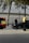 A person in dark clothing, possibly a security or law enforcement officer, is standing next to a black car parked by the roadside. A bright red and yellow vehicle, possibly an emergency service vehicle, is visible nearby. Trees and a metal fence are in the background.