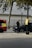 A person in dark clothing, possibly a security or law enforcement officer, is standing next to a black car parked by the roadside. A bright red and yellow vehicle, possibly an emergency service vehicle, is visible nearby. Trees and a metal fence are in the background.
