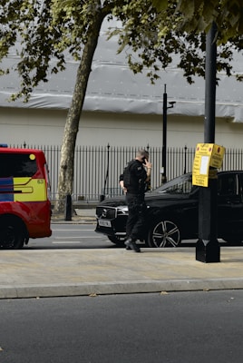 A person in dark clothing, possibly a security or law enforcement officer, is standing next to a black car parked by the roadside. A bright red and yellow vehicle, possibly an emergency service vehicle, is visible nearby. Trees and a metal fence are in the background.