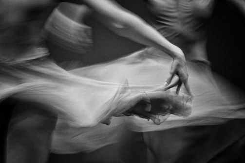 A black and white image capturing the fluid movement of ballet dancers. The focus is on the elegant motion and grace of their arms and fabric, conveying a sense of poetic fluidity and beauty.