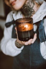 A person holds a lit, dark-colored candle with both hands. The candle is contained in a glass or ceramic holder and emits a warm, flickering light. The person's attire includes a cozy, fleece-like top and overalls.
