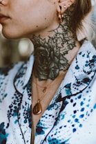 A close-up of a person&rsquo;s neck and part of their face, showcasing a detailed floral tattoo that extends from the neck upwards. Several gold earrings adorn the ear, and the person is wearing a gold necklace with a pendant. The clothing features a pattern in blue and black against a white background.