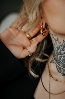 A close-up of a person wearing a gold hoop earring and several rings on their fingers. Visible are a tattoo of a flower on the neck and multiple layered gold necklaces. The person's hand is touching their ear.