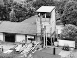 A narrow, multi-story building constructed from brick and concrete stands amidst lush trees. The building features a rooftop terrace with a banner and power lines running across the scene. Several people are working on roofing materials or walking on the street nearby.