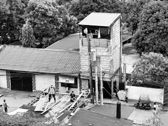 A narrow, multi-story building constructed from brick and concrete stands amidst lush trees. The building features a rooftop terrace with a banner and power lines running across the scene. Several people are working on roofing materials or walking on the street nearby.