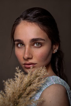 A young person with expressive eyes and natural makeup is holding a bunch of dried grasses. They are wearing a light blue and white striped sleeveless top. The background is a muted, dark color, accentuating the subject's features.