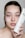 A woman holds a tube of skincare product close to her face. She has a patch of a creamy pink mask applied on her cheek and is looking directly at the camera. Her nails feature delicate black and white designs.