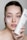 A woman holds a tube of skincare product close to her face. She has a patch of a creamy pink mask applied on her cheek and is looking directly at the camera. Her nails feature delicate black and white designs.