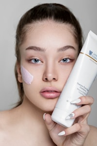 A woman holds a tube of skincare product close to her face. She has a patch of a creamy pink mask applied on her cheek and is looking directly at the camera. Her nails feature delicate black and white designs.