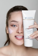 A person with light skin is smiling and holding up a tube of Janssen Cosmetics Thermo Peel Mask Cranberry next to their face. They have applied some of the mask to their cheek, and their nails have a delicate design.