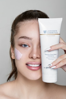 A person with light skin is smiling and holding up a tube of Janssen Cosmetics Thermo Peel Mask Cranberry next to their face. They have applied some of the mask to their cheek, and their nails have a delicate design.