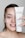 A person with light skin is smiling and holding up a tube of Janssen Cosmetics Thermo Peel Mask Cranberry next to their face. They have applied some of the mask to their cheek, and their nails have a delicate design.