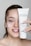 A person with light skin is smiling and holding up a tube of Janssen Cosmetics Thermo Peel Mask Cranberry next to their face. They have applied some of the mask to their cheek, and their nails have a delicate design.
