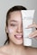 A person with light skin is smiling and holding up a tube of Janssen Cosmetics Thermo Peel Mask Cranberry next to their face. They have applied some of the mask to their cheek, and their nails have a delicate design.