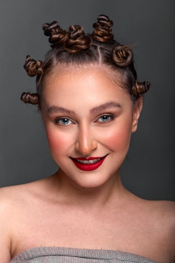 A person with Bantu knots styled in their hair, wearing bold red lipstick and natural makeup. The background is a neutral gray, and they are smiling warmly.