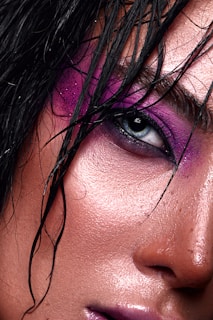 A close-up of an eye with dramatic purple and pink eye makeup. The skin is smooth and glistening, with strands of wet hair falling across the face.