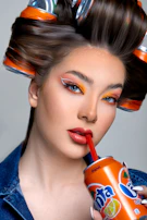 A woman with elaborate makeup, featuring bold orange and red tones, is sipping from a Fanta can using a red straw. Her hair is styled with soda cans as curlers, blending fashion with unconventional elements.