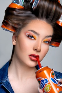 A woman with elaborate makeup, featuring bold orange and red tones, is sipping from a Fanta can using a red straw. Her hair is styled with soda cans as curlers, blending fashion with unconventional elements.