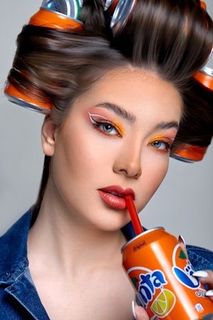 A woman with elaborate makeup, featuring bold orange and red tones, is sipping from a Fanta can using a red straw. Her hair is styled with soda cans as curlers, blending fashion with unconventional elements.