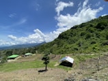 A rural landscape featuring a hilly terrain covered with lush green trees and vegetation. In the foreground, there are small structures with green roofs, and a makeshift shelter with a metal roof. The sky is bright and clear with scattered fluffy clouds.