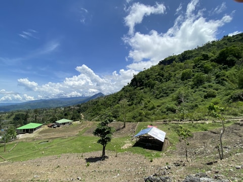 A rural landscape featuring a hilly terrain covered with lush green trees and vegetation. In the foreground, there are small structures with green roofs, and a makeshift shelter with a metal roof. The sky is bright and clear with scattered fluffy clouds.