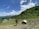 A rural landscape featuring a hilly terrain covered with lush green trees and vegetation. In the foreground, there are small structures with green roofs, and a makeshift shelter with a metal roof. The sky is bright and clear with scattered fluffy clouds.