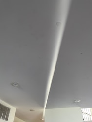 A modern interior ceiling with recessed lights and a light-colored surface. A subtle curved design is visible, along with an air vent on the left side.