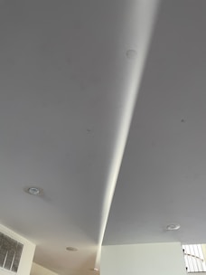 A modern interior ceiling with recessed lights and a light-colored surface. A subtle curved design is visible, along with an air vent on the left side.