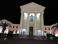 A grand neoclassical building with tall columns and a gable roof, well-lit against a dark night sky. The front entrance features large wooden doors, above which are Russian letters. Palm trees flank the front of the building, and a few people are walking nearby.
