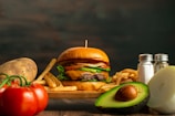 A juicy hamburger with lettuce, tomato, and cheese sits on a wooden board surrounded by crispy French fries. Fresh ingredients such as a whole avocado, sliced onion, a potato, and ripe tomatoes accompany the scene. A pair of salt and pepper shakers is positioned on the right side.