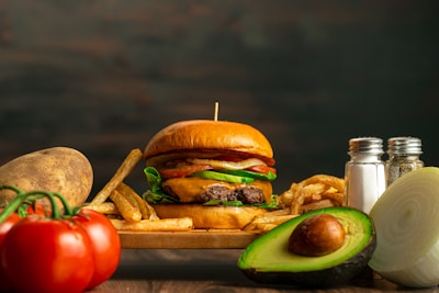 A juicy hamburger with lettuce, tomato, and cheese sits on a wooden board surrounded by crispy French fries. Fresh ingredients such as a whole avocado, sliced onion, a potato, and ripe tomatoes accompany the scene. A pair of salt and pepper shakers is positioned on the right side.