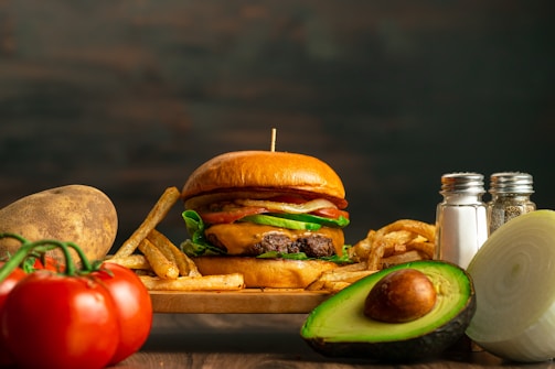 A juicy hamburger with lettuce, tomato, and cheese sits on a wooden board surrounded by crispy French fries. Fresh ingredients such as a whole avocado, sliced onion, a potato, and ripe tomatoes accompany the scene. A pair of salt and pepper shakers is positioned on the right side.